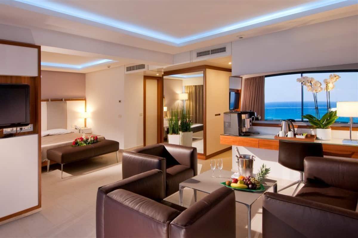 Habitación de hotel moderna con zona de estar, una mesa de comedor con fruta, televisor y vista al océano.