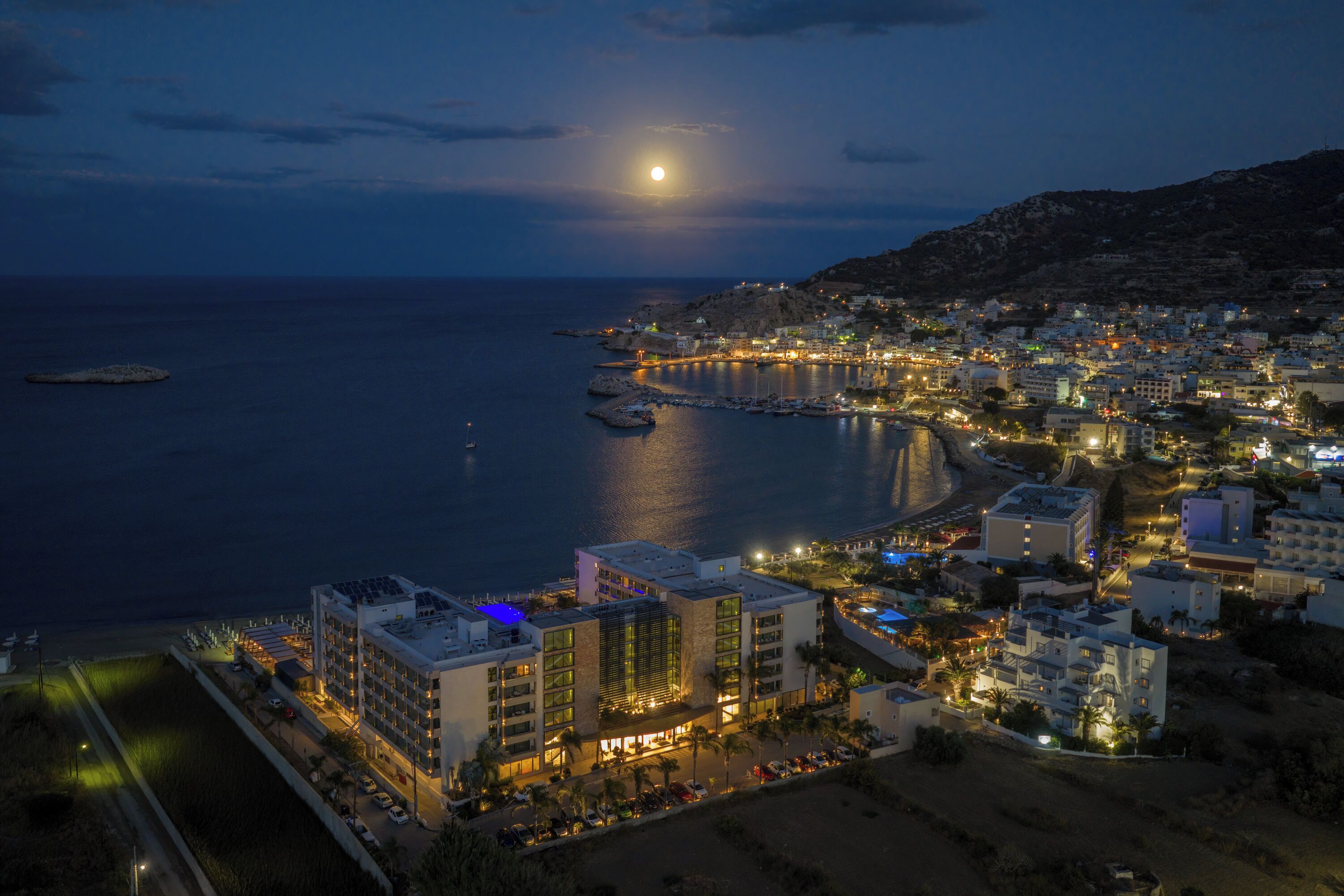 A coastal cityscape at night with a prominent building featuring multiple floors and balconies, a marina with boats, and a beach area.