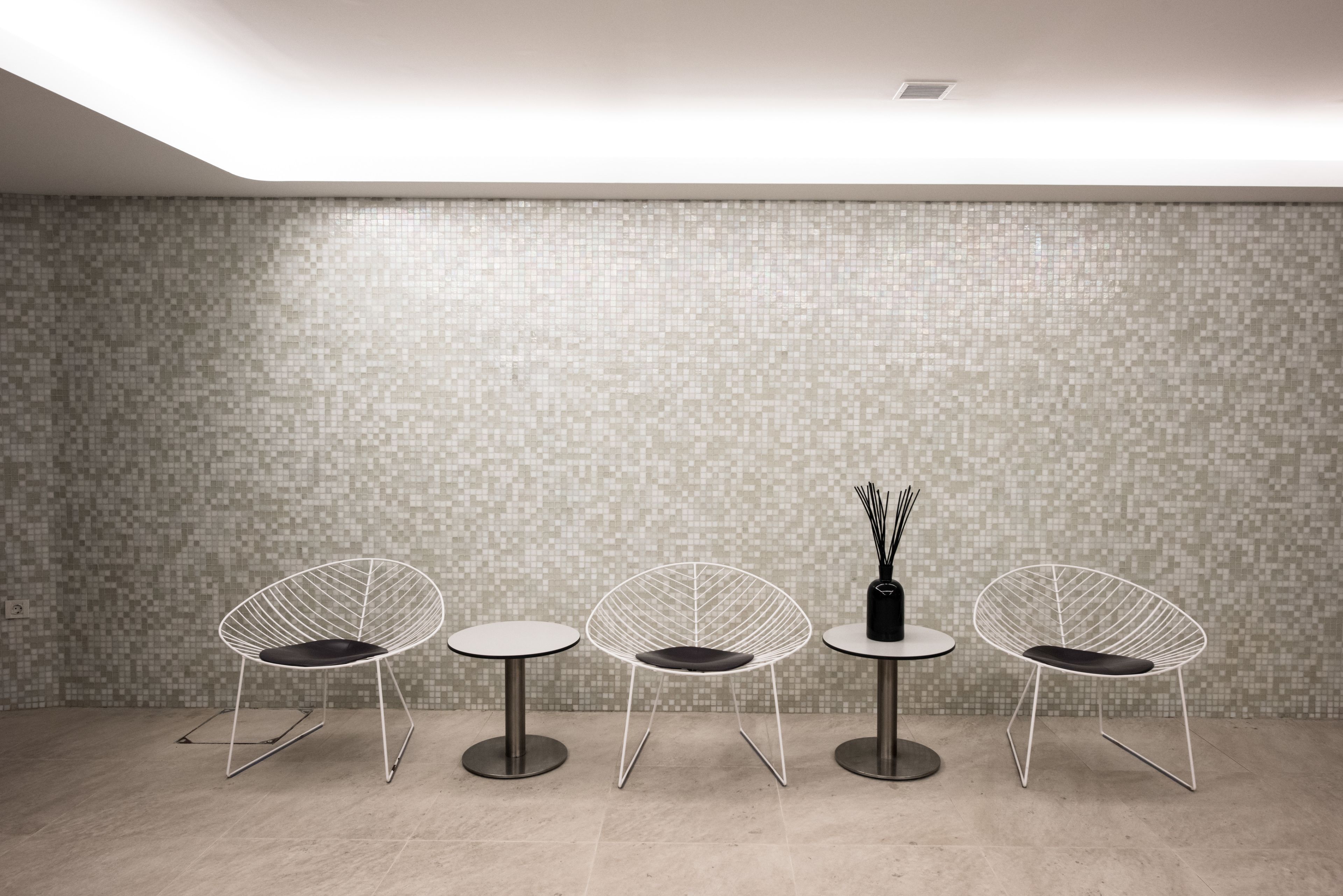 Un espace d’attente moderne avec trois chaises blanches à structure métallique et deux tables rondes, dont l’une est ornée d’un vase noir et d’une paire de bâtonnets d’encens.