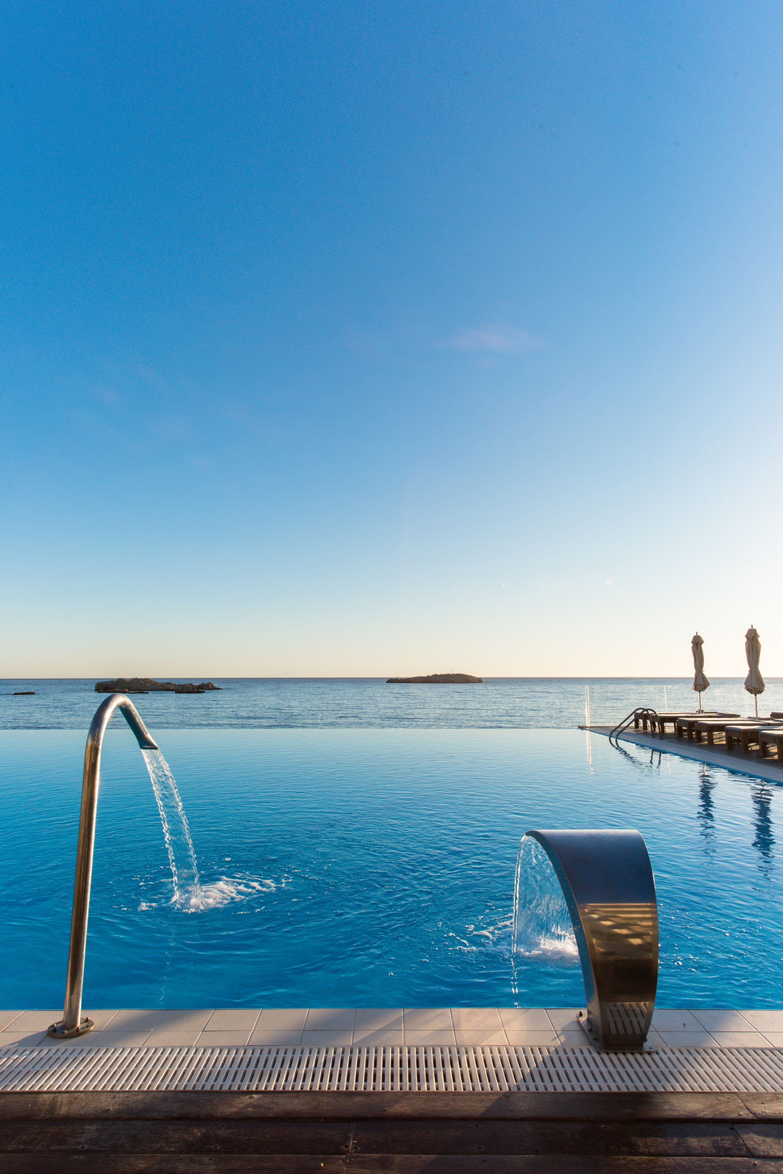 An infinity pool with a clear view of the ocean, a water fountain, and a deck with umbrellas.