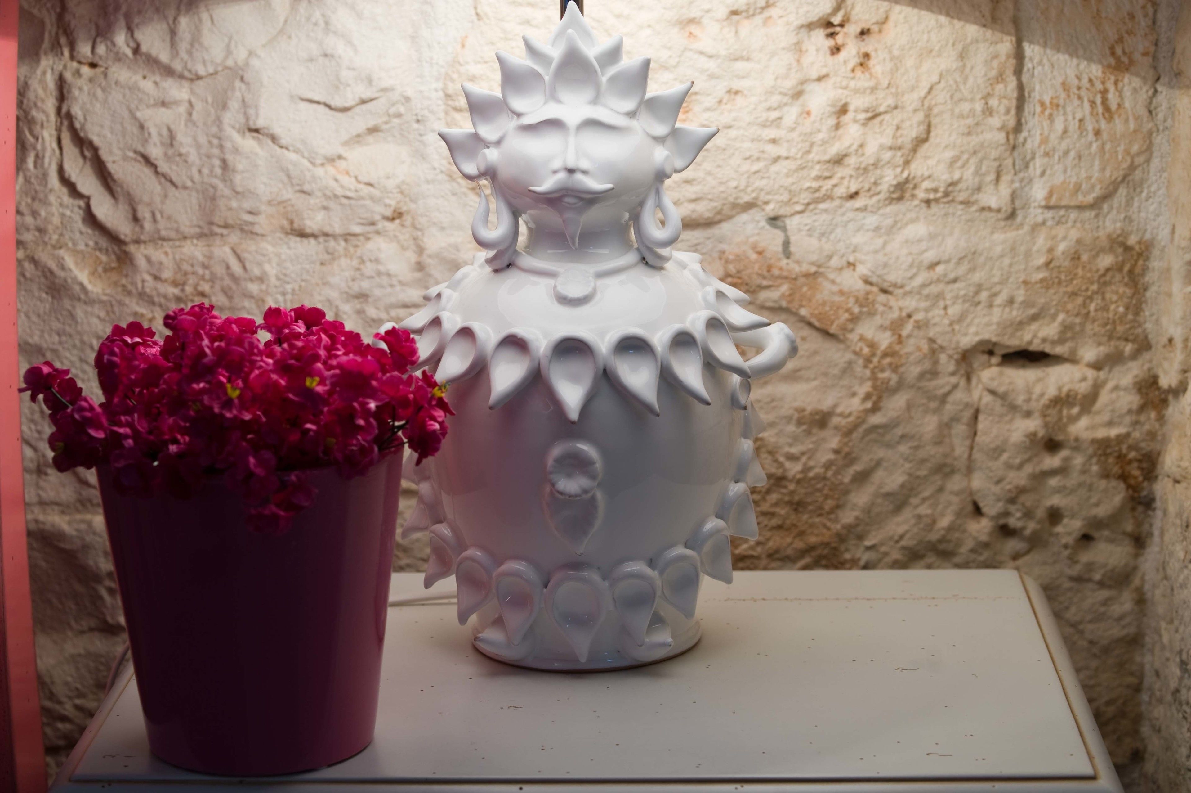 Uma escultura de cerâmica branca com rosto e um vaso com flores cor-de-rosa sobre uma superfície branca.