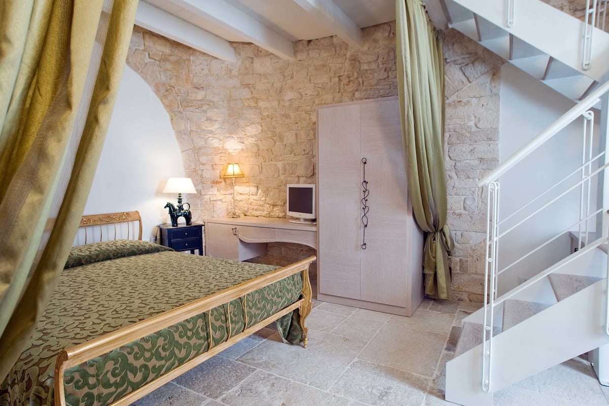 Une chambre avec un mur en pierre, un lit, un bureau avec une lampe, une armoire et un escalier.