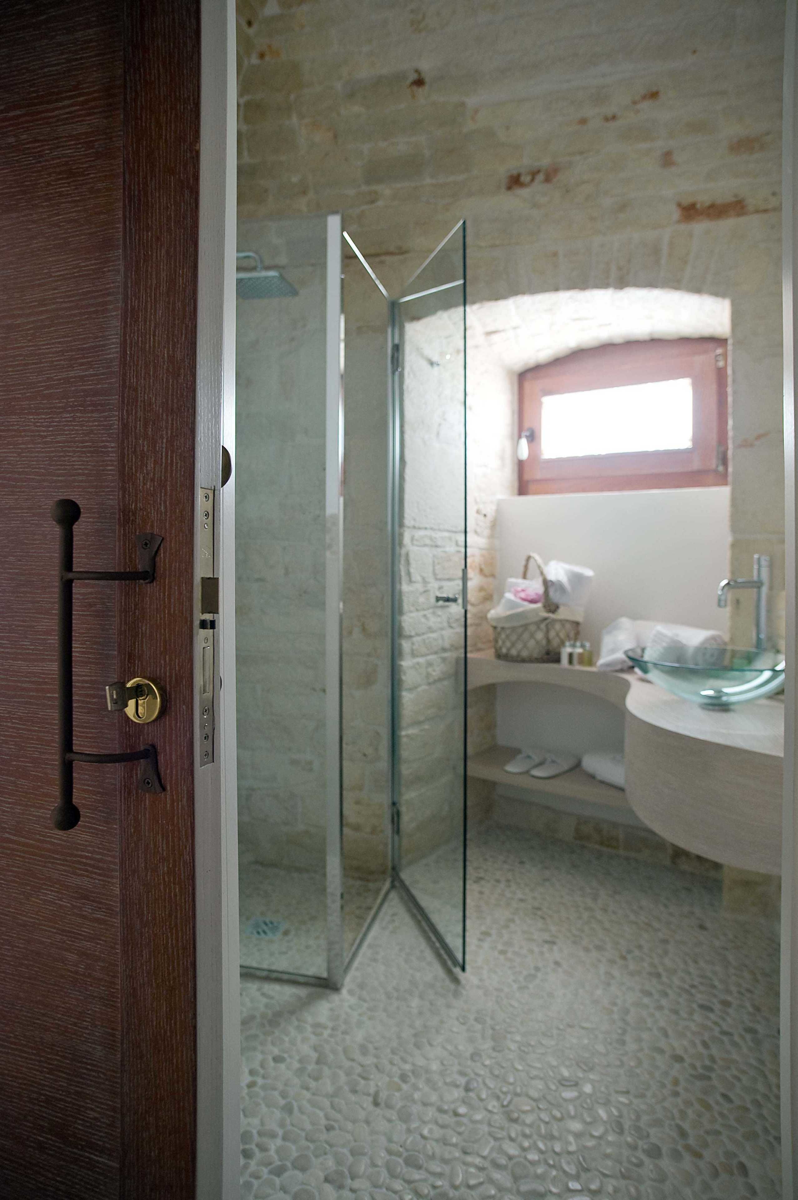 Ein Badezimmer mit Duschkabine aus Glas, einer Holztür, Steinböden, einem weißen Waschbecken und einem Fenster über dem Waschbecken.