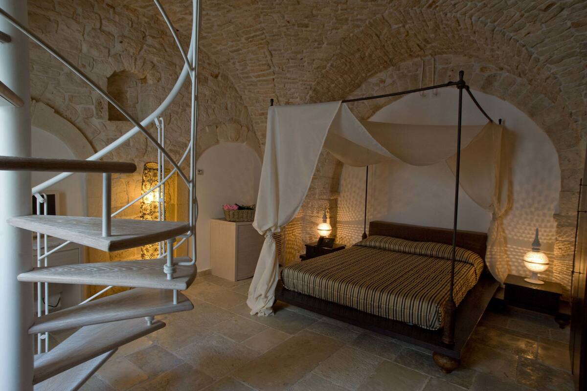 Une chambre avec un plafond en pierre, un lit à baldaquin, un escalier et une table de chevet avec une lampe.
