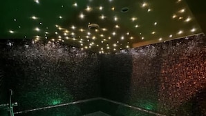 A spa room with a green-lit jacuzzi, dark tiled walls, and numerous ceiling lights.