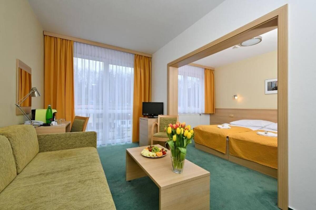 Une chambre d’hôtel avec un canapé, une petite table sur laquelle se trouve une assiette de fruits, un vase avec des fleurs, un lit et un téléviseur.