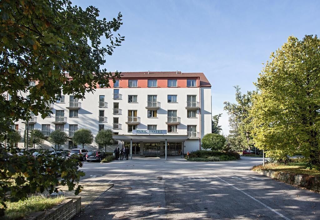 Un hotel de varios pisos con una entrada central, rodeado de árboles y automóviles estacionados.