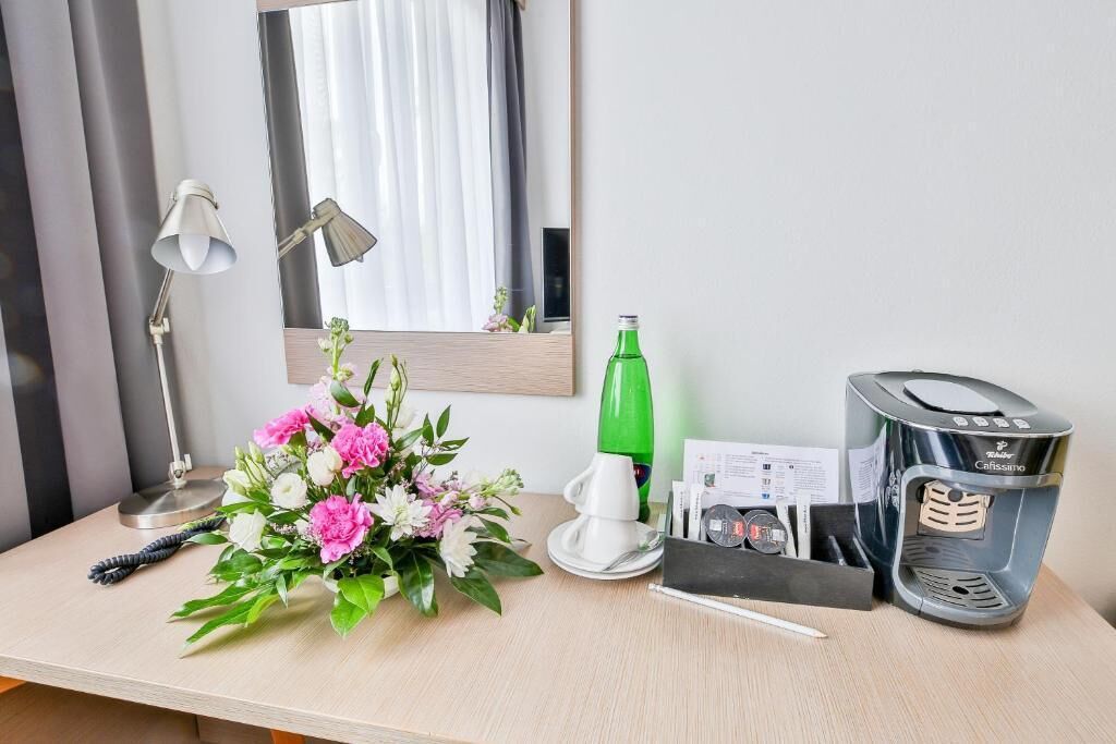 Ett skrivbord i ett hotellrum med en lampa, en blomsterbukett, en grön flaska, en kaffemaskin och ett anteckningsblock.