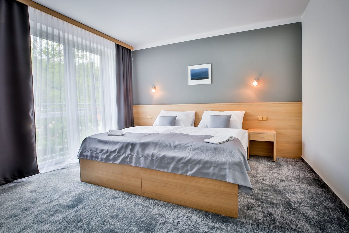 Une chambre d’hôtel moderne avec un grand lit, une tête de lit en bois, deux tables de chevet et une vue sur la verdure à travers des rideaux transparents.
