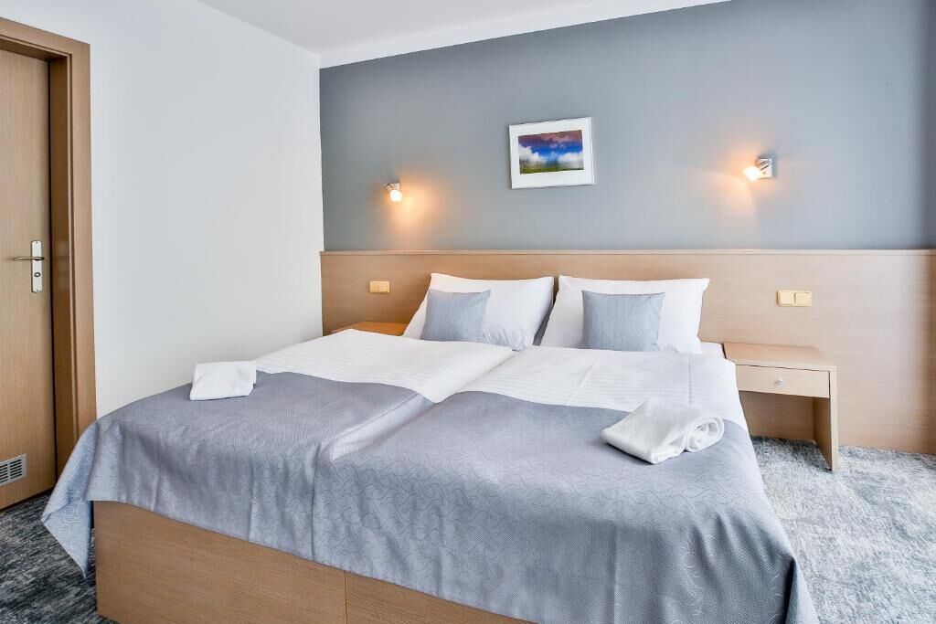 Un lit double avec une couvre-lit grise et des oreillers blancs, deux tables de chevet avec des lampes, une porte en bois et un tableau encadré au mur.