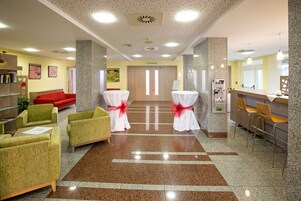 Un vestíbulo de hotel con una mesa de recepción, un sofá rojo, sillones verdes y piso alfombrado.