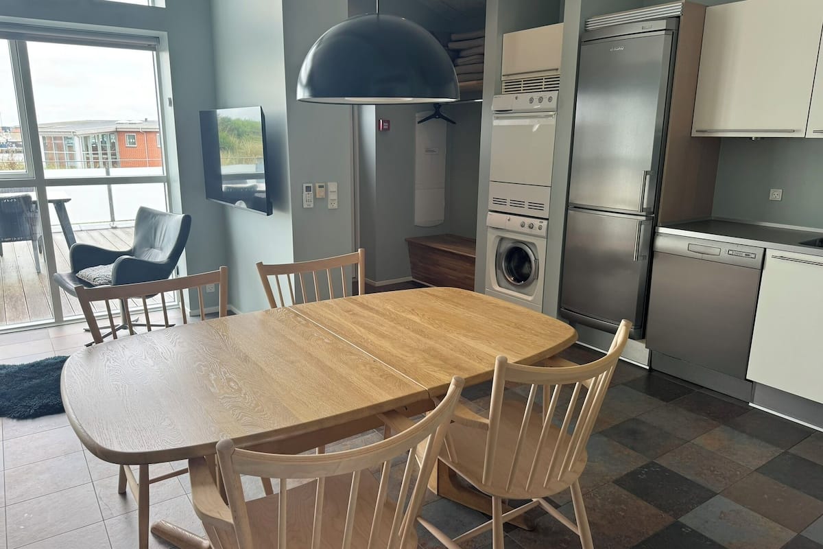 Una cocina moderna con una mesa y sillas de madera, lavadora y secadora integradas, y un amplio ventanal con vistas a una terraza.