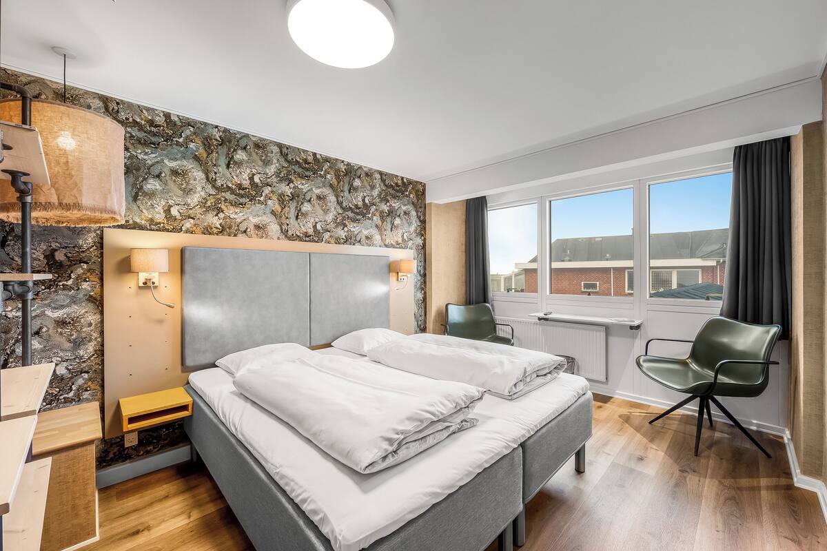 Habitación de hotel moderna con pared de acento de piedra, una cama grande con ropa blanca, un escritorio con una silla y una ventana con vistas a edificios.