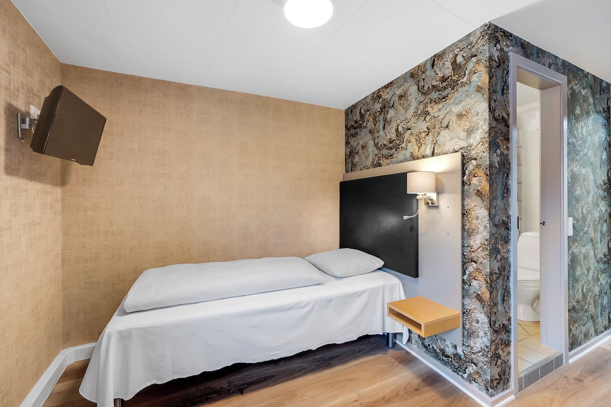 Un dormitorio con cama, mesita de noche, televisor montado en la pared y baño visible a través de un separador de vidrio.