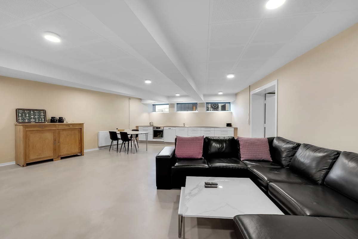 Una sala de estar moderna con un sofá de cuero negro, una mesa de centro blanca y un aparador de madera. Se observa una cocina al fondo con armarios blancos y un comedor con una mesa y sillas.
