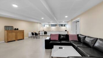Una sala de estar moderna con un sofá de cuero negro, una mesa de centro blanca y un aparador de madera. Se observa una cocina al fondo con armarios blancos y un comedor con una mesa y sillas.