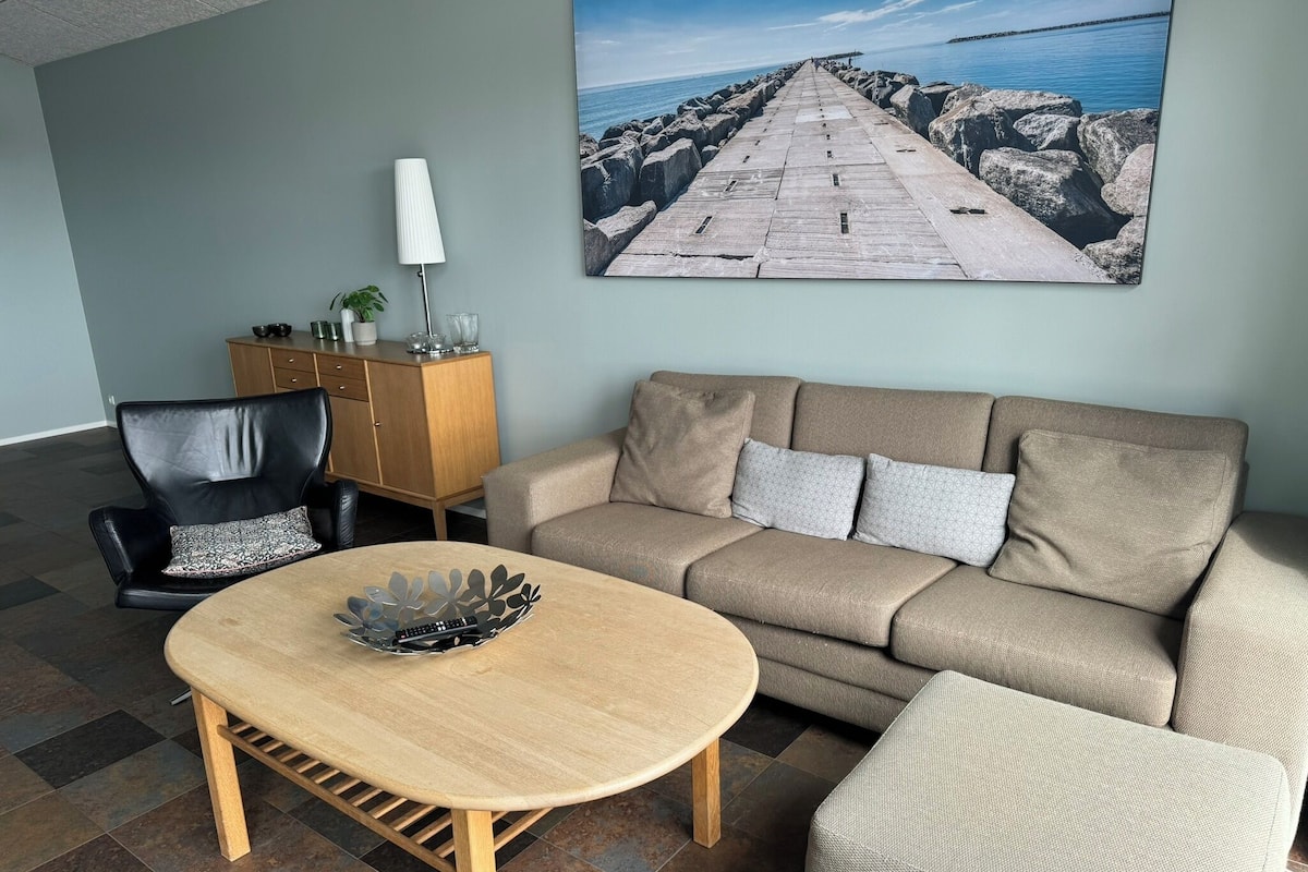 Una sala de estar con un sofá beige, una mesa de centro de madera, un sillón de cuero negro y un aparador. En la pared, un cuadro grande de un muelle y agua.