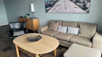 Una sala de estar con un sofá beige, una mesa de centro de madera, un sillón de cuero negro y un aparador. En la pared, un cuadro grande de un muelle y agua.