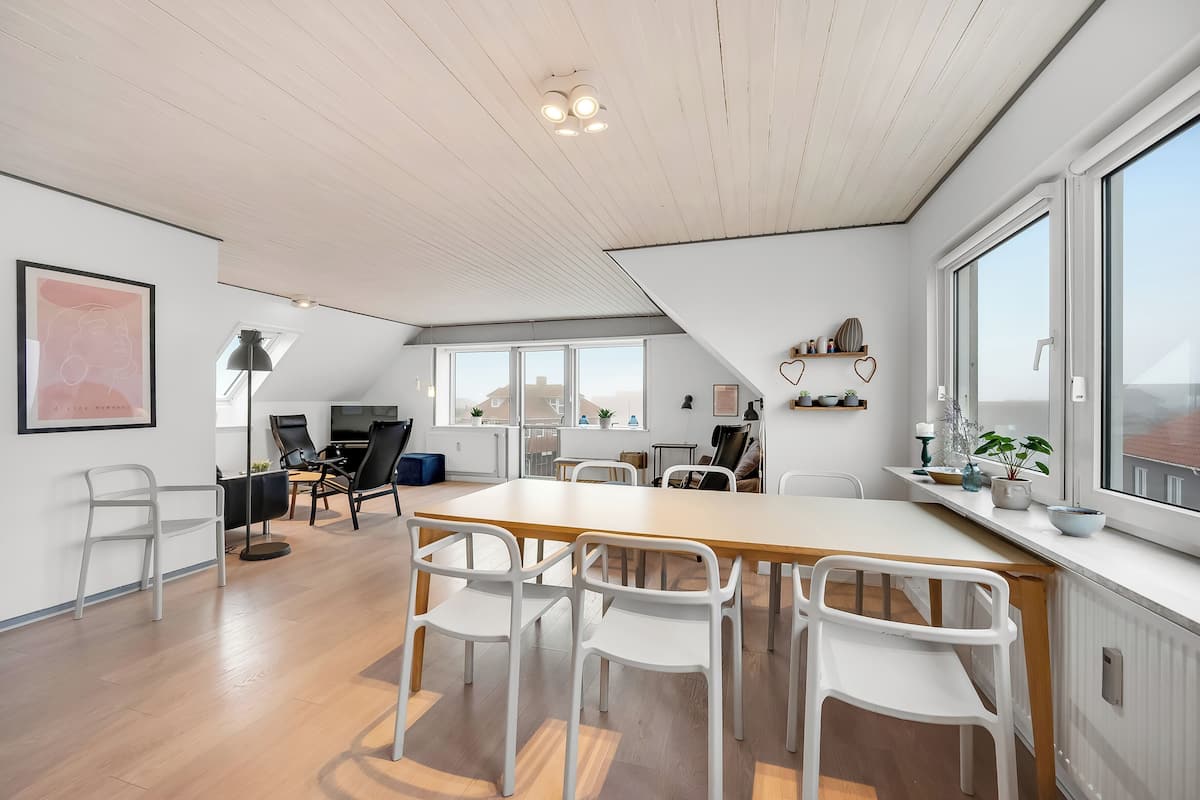 Una habitación amplia con una gran mesa de madera, sillas blancas y vistas amplias a través de ventanas grandes.