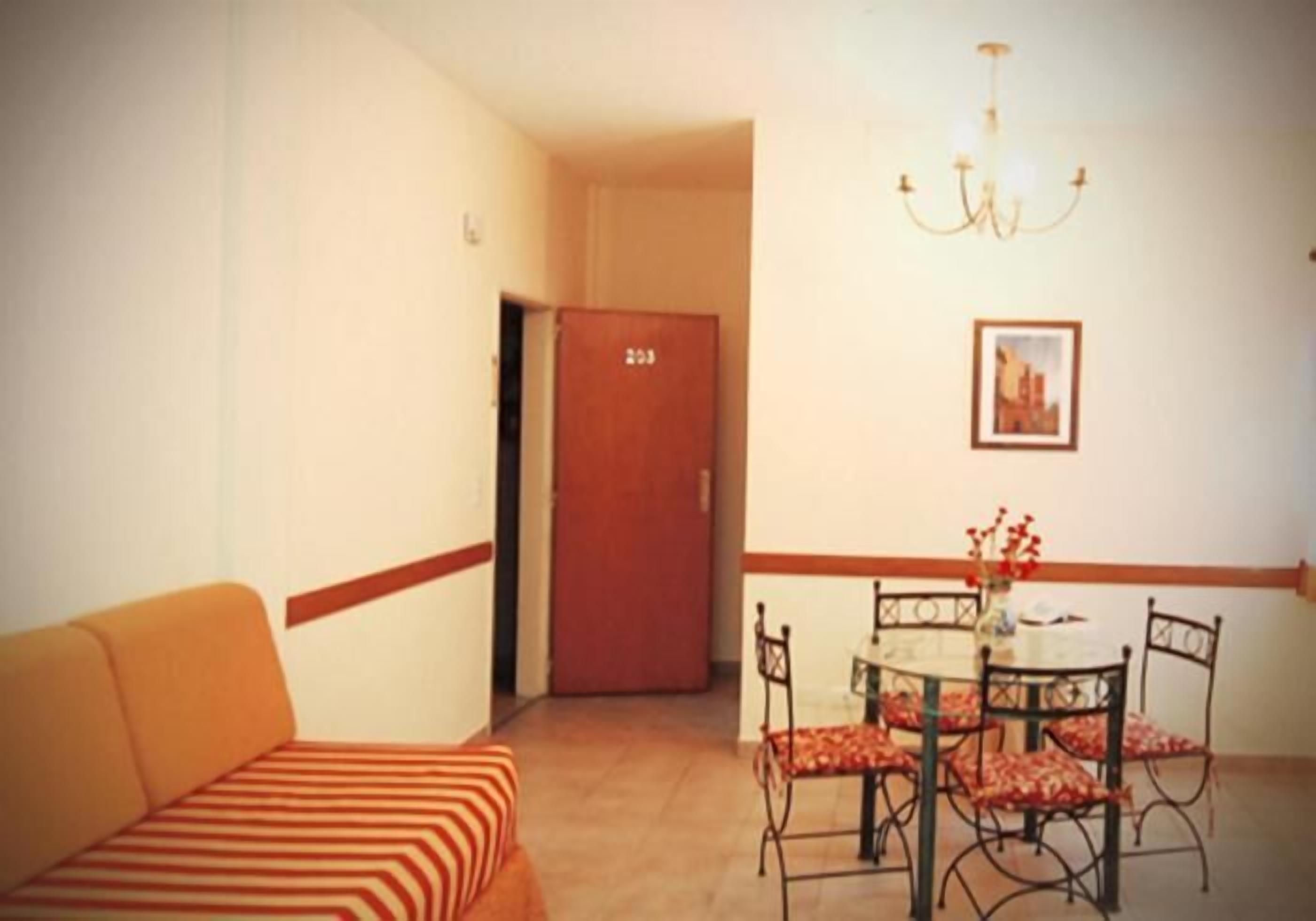 A room with a sofa, a dining table with chairs, a chandelier, and a door with a number.