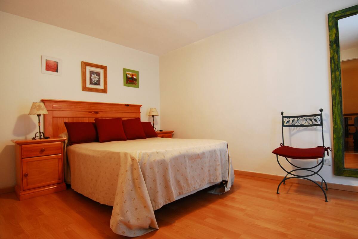 Ein Schlafzimmer mit einem hölzernen Bett, zwei Nachttischen mit Lampen, einem roten Sessel und einem Spiegel an der Wand.