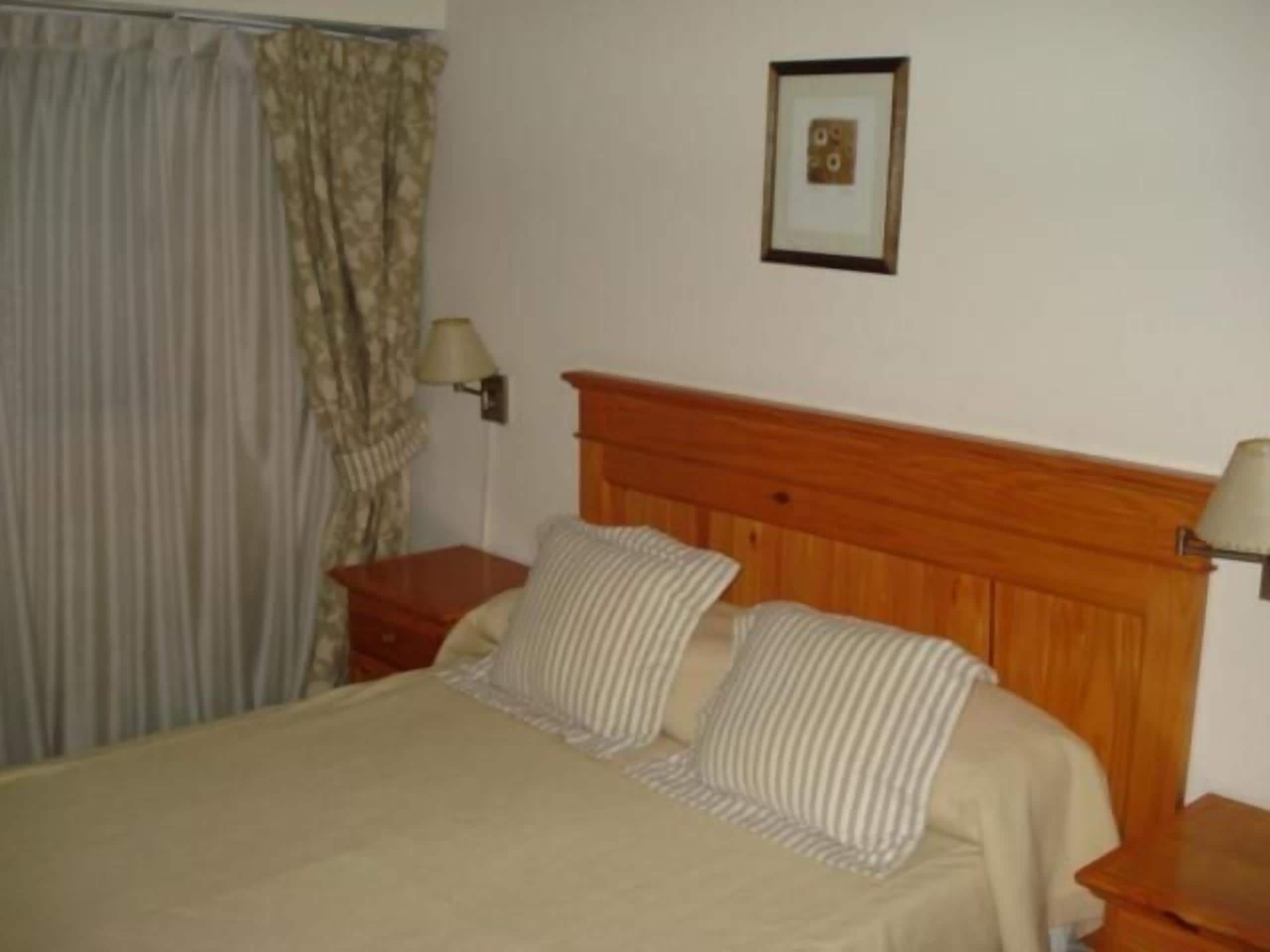Een netjes opgemaakt bed met een houten hoofdbord, een nachtkastje met een lamp, en een gordijn dat deels is opengetrokken.