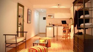 Un espace de vie compact comprenant un coin cuisine, un coin repas avec des chaises à rayures, un lit avec un cadre en métal et une armoire avec des étagères.