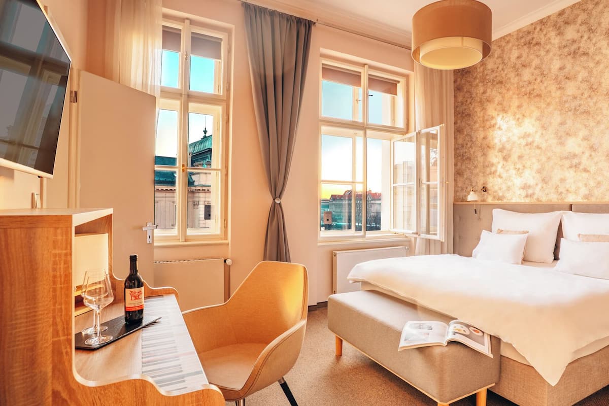 Een hotelkamer met een bed, een bureau met een fles wijn en glas, een stoel, en uitzicht op gebouwen door grote ramen.