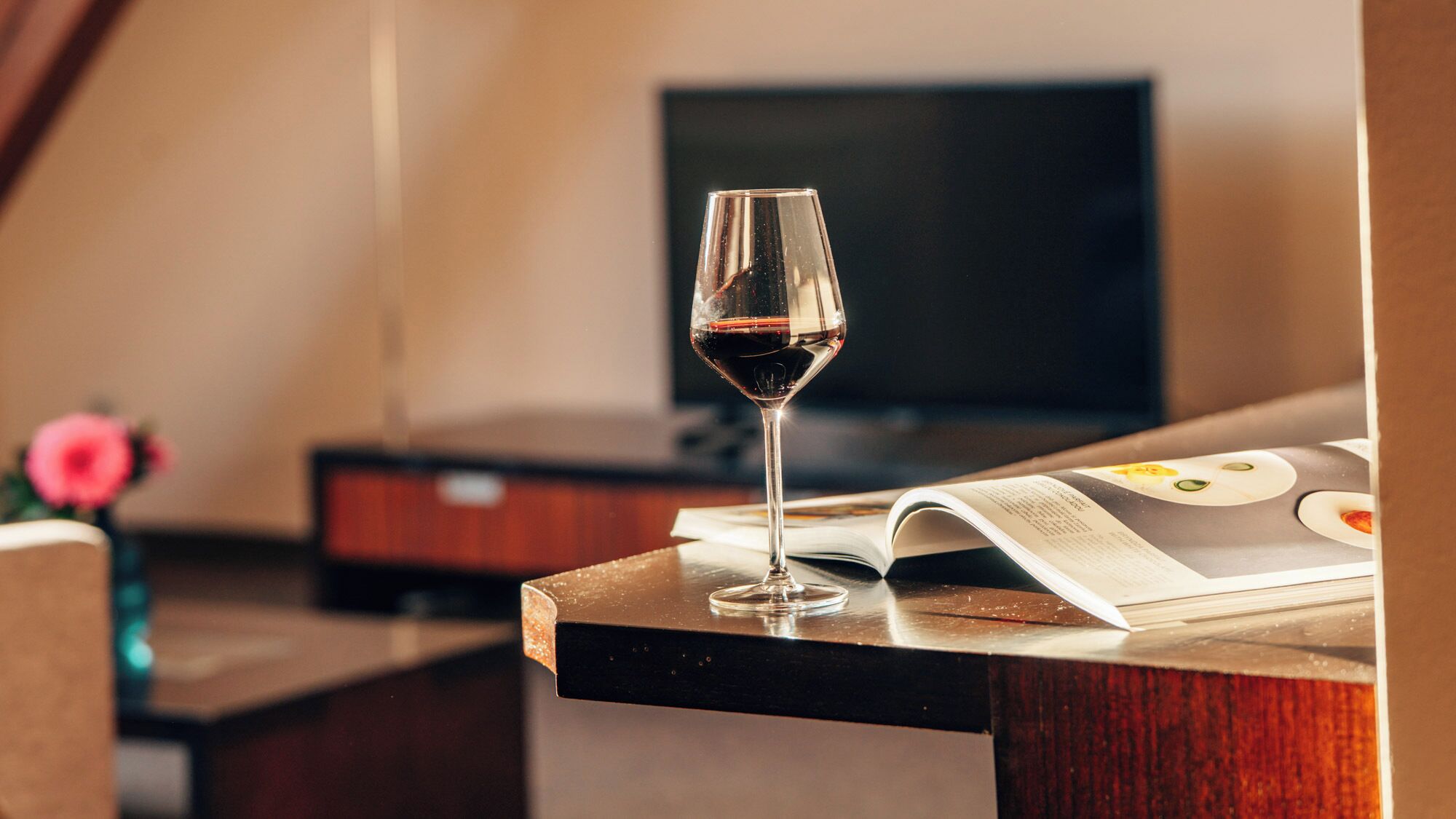 Ein Glas Rotwein auf einer Holztischplatte mit einer Zeitschrift, einem Fernseher im Hintergrund und einer rosa Blume auf der linken Seite.