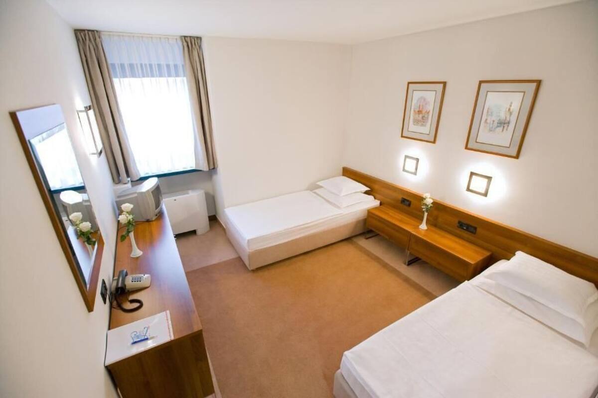 A hotel room with a single bed, a smaller bed, a desk with a telephone, a mirror, and a window with curtains.