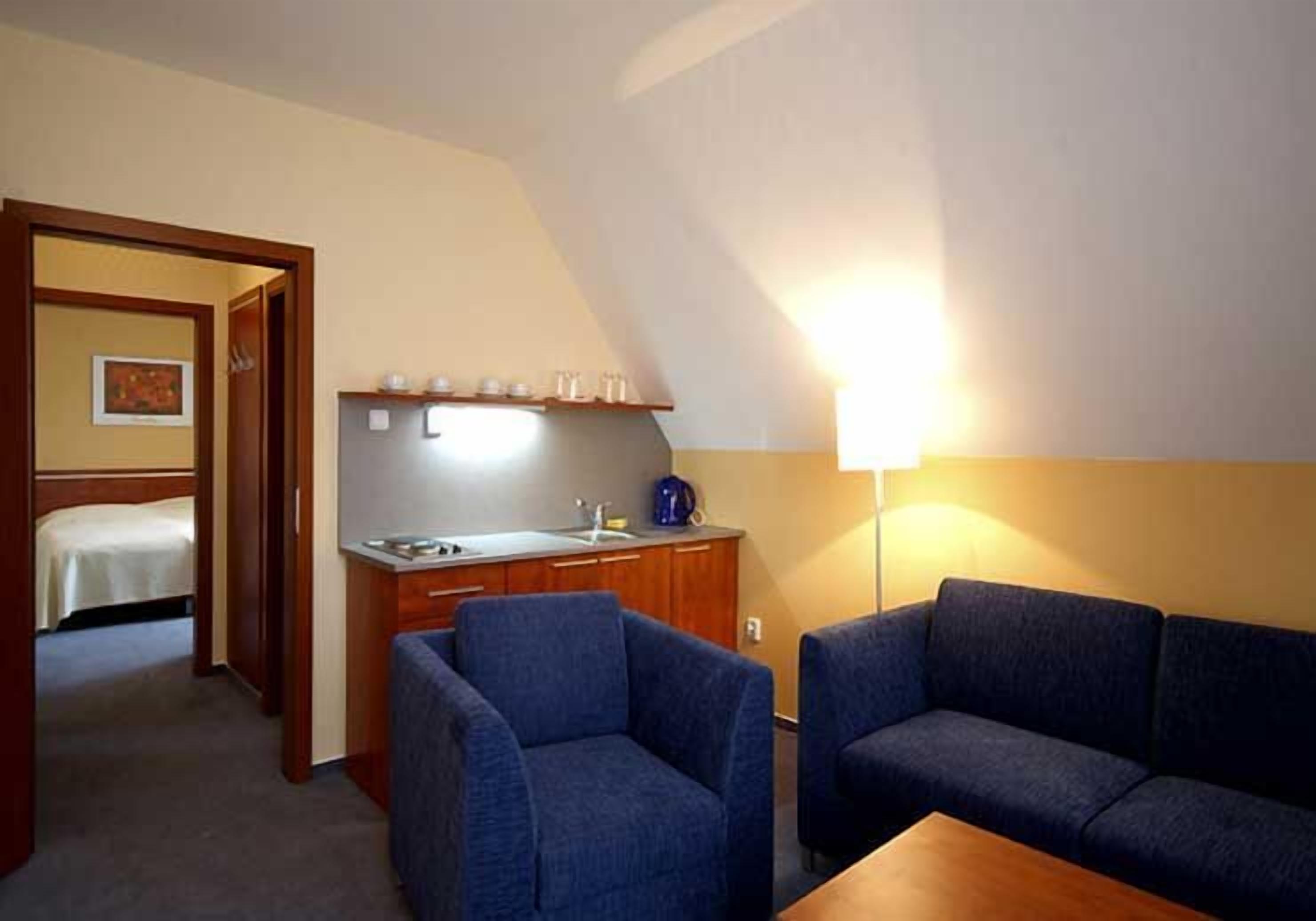 A compact room with a kitchenette, a bed, and two blue armchairs.