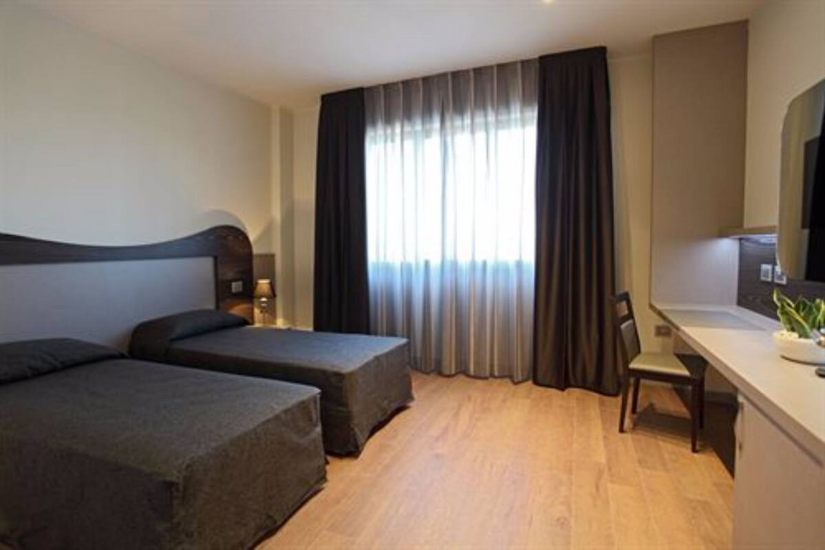 A hotel room with two single beds, a desk with a chair, a TV, and a large window with curtains.