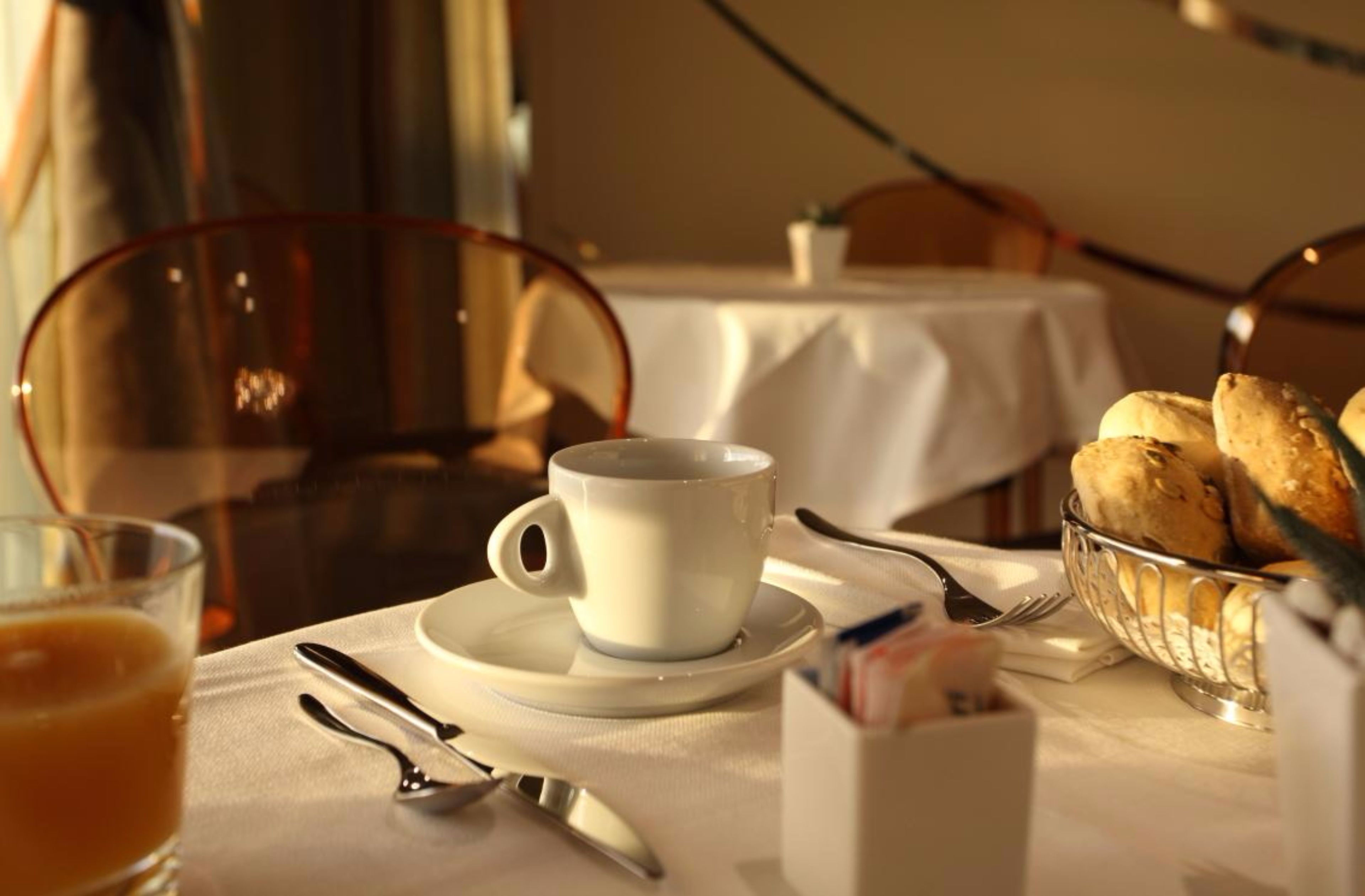 Una mesa con desayuno: una taza, un platillo, utensilios, un vaso de jugo de naranja y una cesta de pasteles.