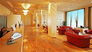 A hotel reception area with a polished wooden floor, red leather sofas, a reception desk, and a chandelier. - Grand Hotel Forlì (Forlì)
