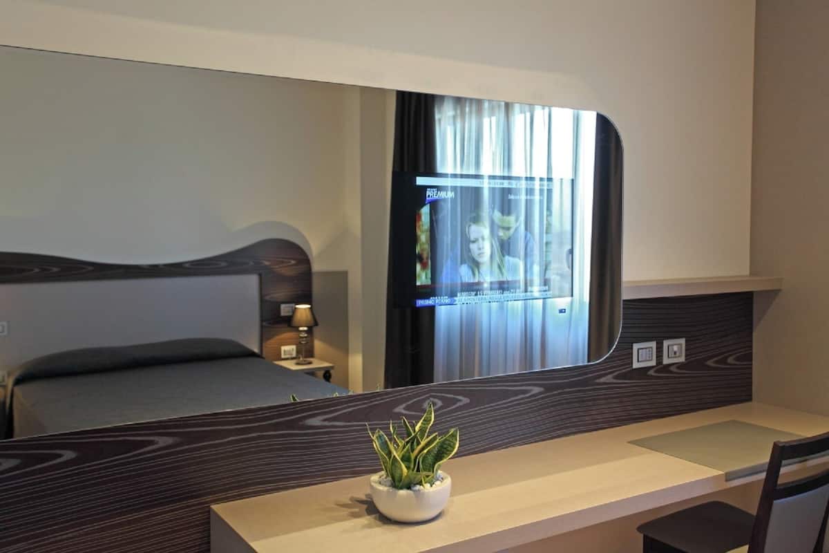 A hotel room with a bed, a bedside table with a lamp, a plant on the table, and a large mirror reflecting the room.