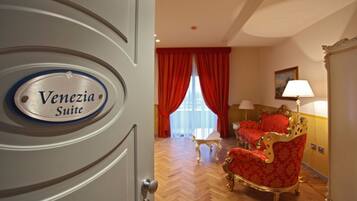 Habitación de hotel con suelo de parqué tipo espiga, una silla roja y dorada ornamentada, una mesa blanca y una lámpara.