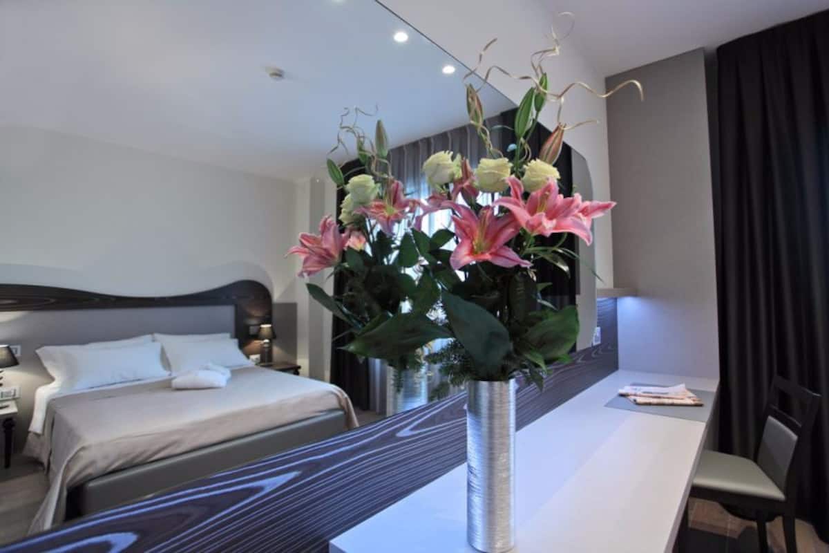 A modern hotel room with a large bed, a desk with a vase of flowers, and a view of the city.