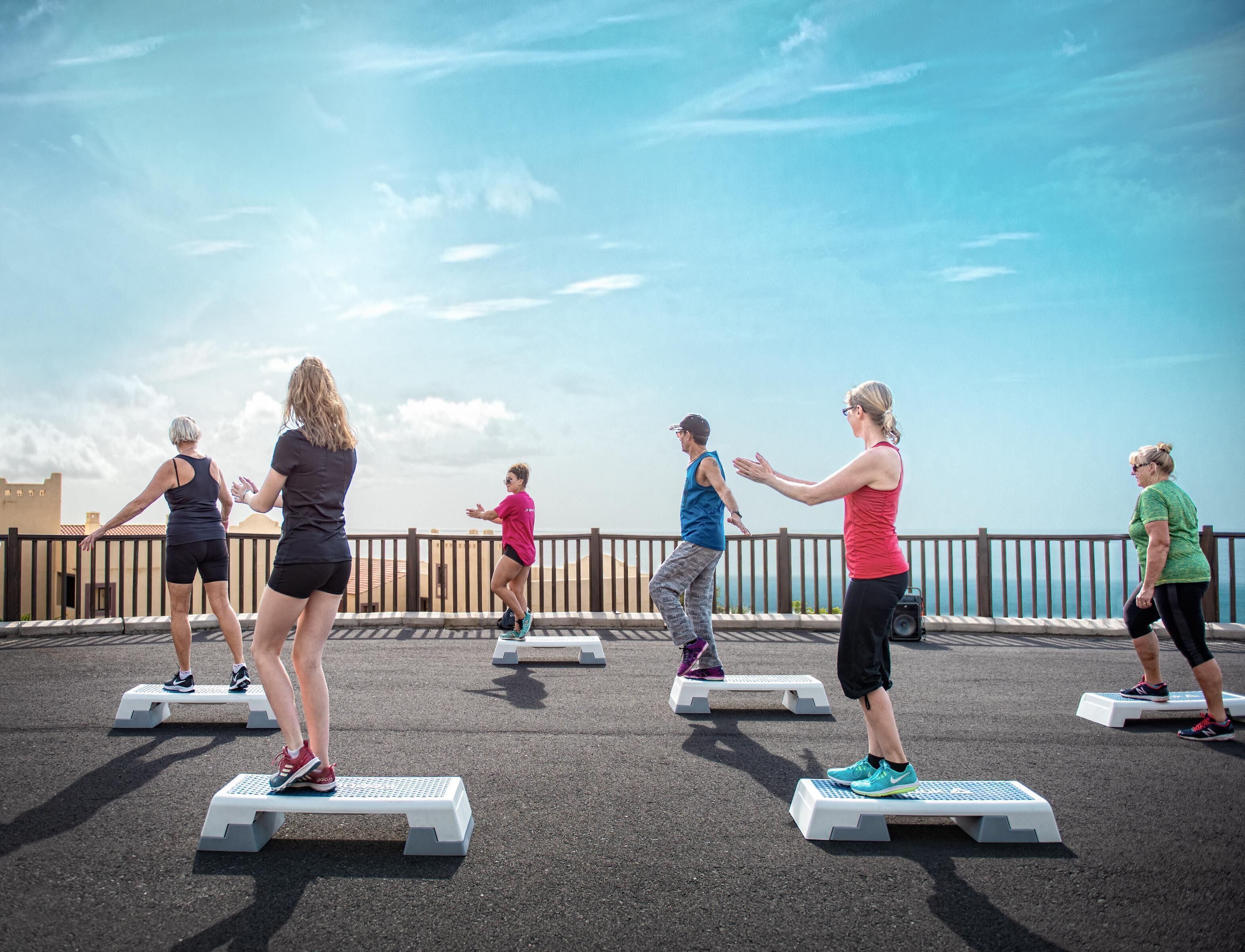A group of people exercising on step platforms outdoors with a clear sky in the background.