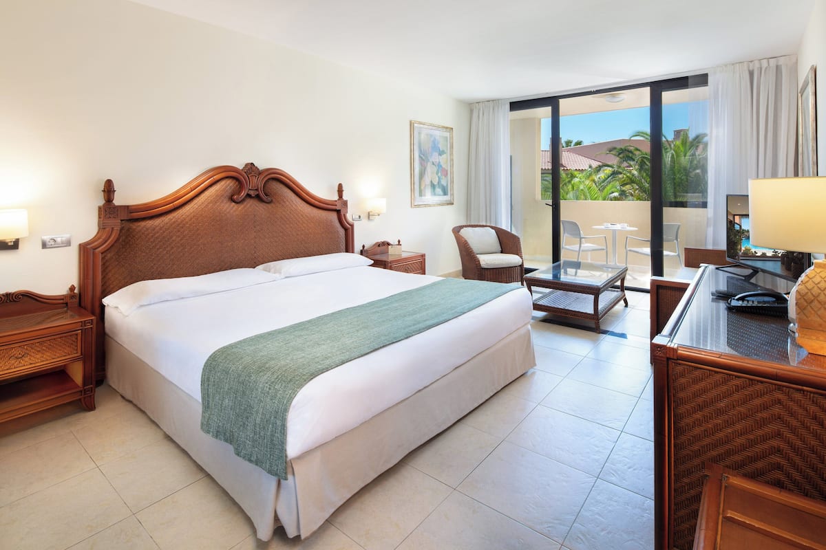 Une chambre d’hôtel avec un grand lit, une chaise, un bureau et un balcon donnant sur des palmiers.