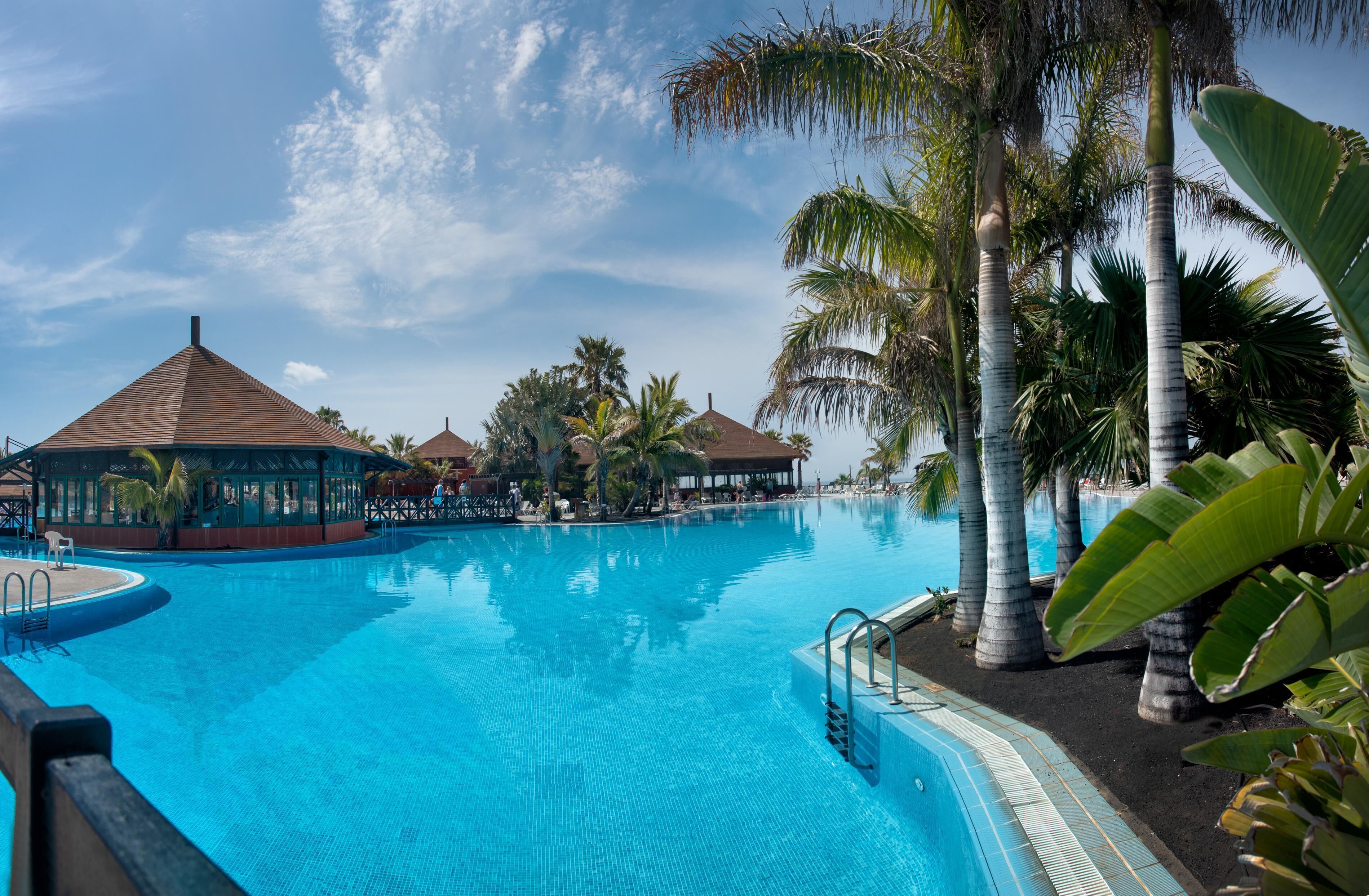 A clear blue pool with palm trees and a gazebo in the background.