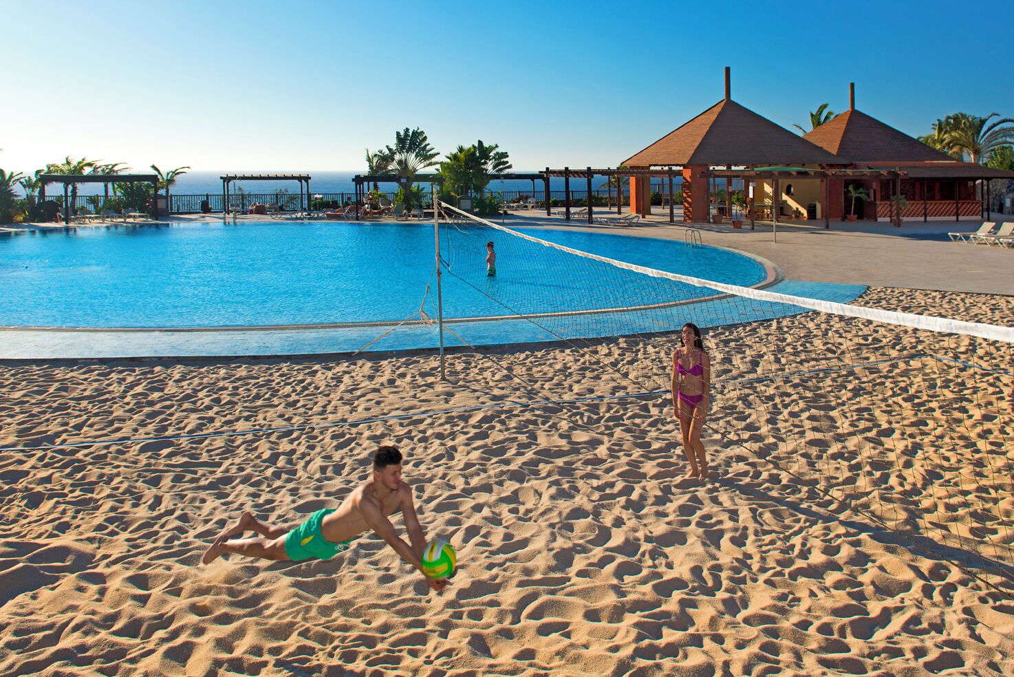 Un terrain de beach-volley situé à côté d’une piscine, offrant une vue dégagée sur l’océan.