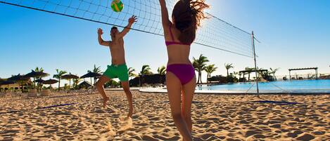 A sandy beach with a volleyball net, palm trees, and a pool in the background.