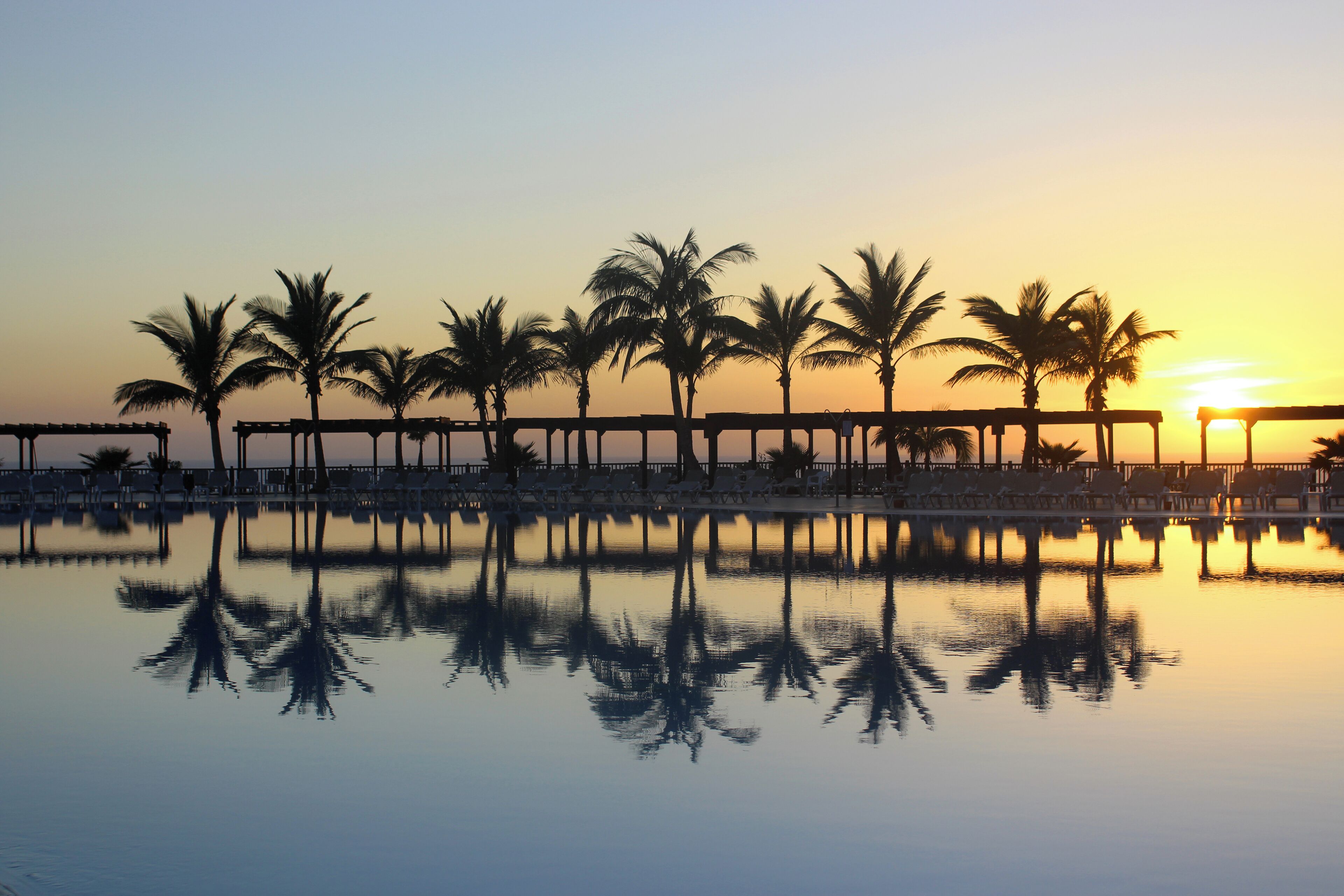 A serene sunset over a calm pool with palm trees and lounge chairs.