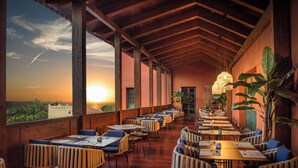A restaurant with a long table layout, large windows, and a view of the sunset. - Hotel La Palma Princess (Fuencaliente de la Palma)