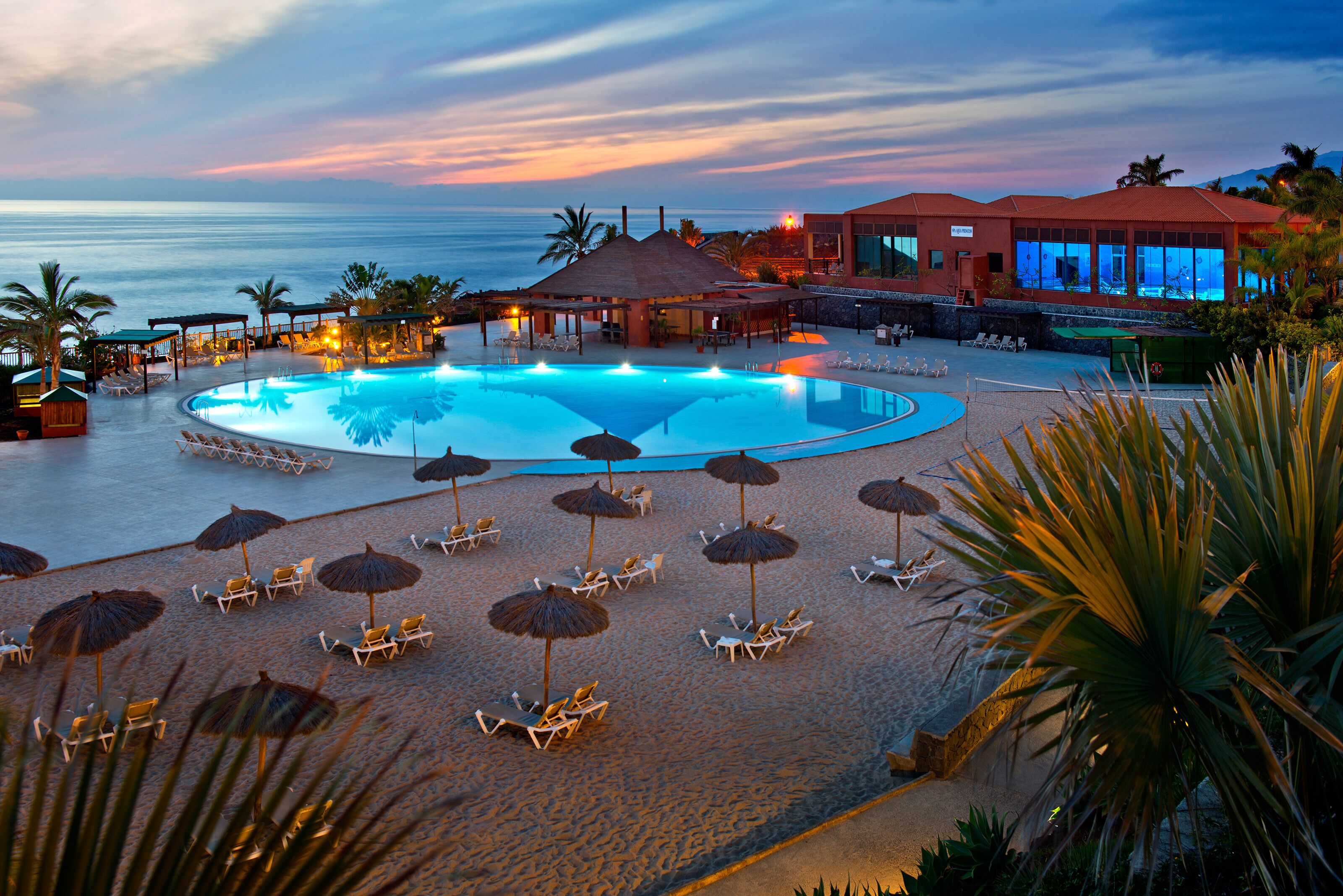 A resort pool area with lounge chairs and umbrellas, surrounded by a building with a red roof.