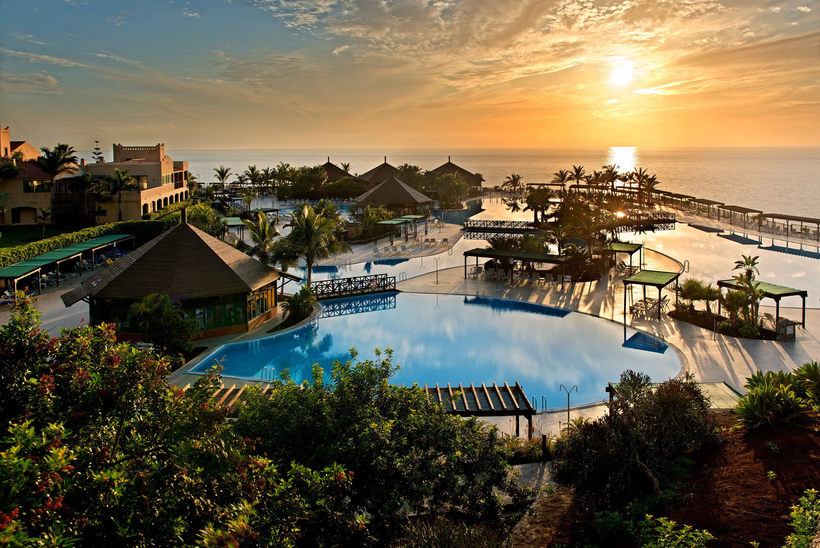 Un resort con una amplia piscina, bungalows sobre el agua y vistas al atardecer.