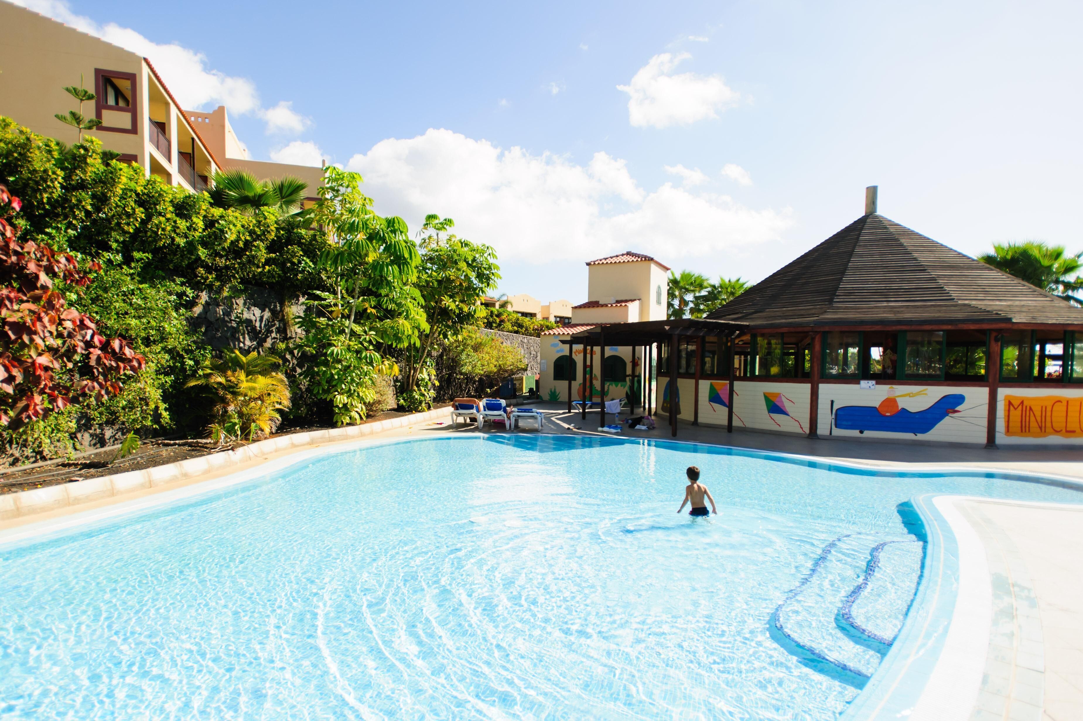 A swimming pool with a child playing, surrounded by greenery and a building with a mural.