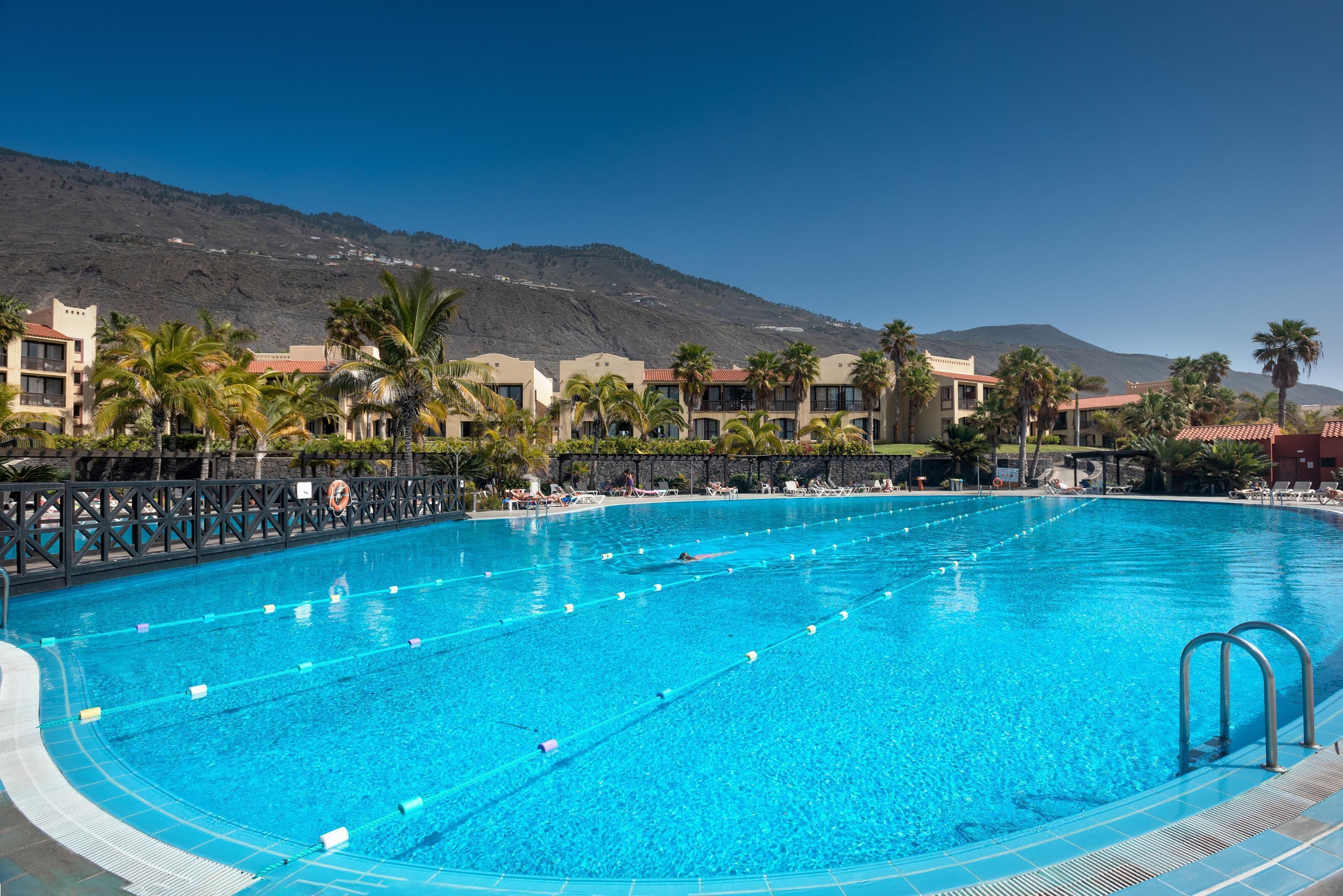 A swimming pool with clear blue water, surrounded by palm trees and a mountain in the background.