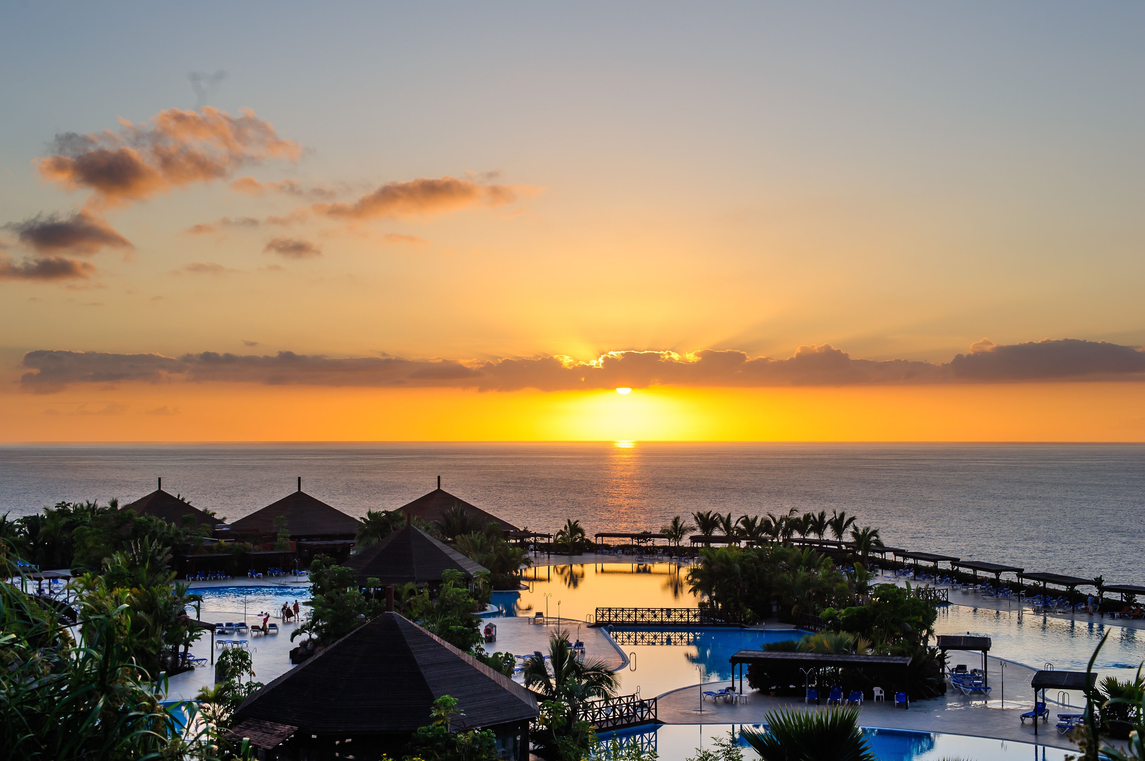A resort with a pool area, lounge chairs, and a view of the sunset over the ocean.
