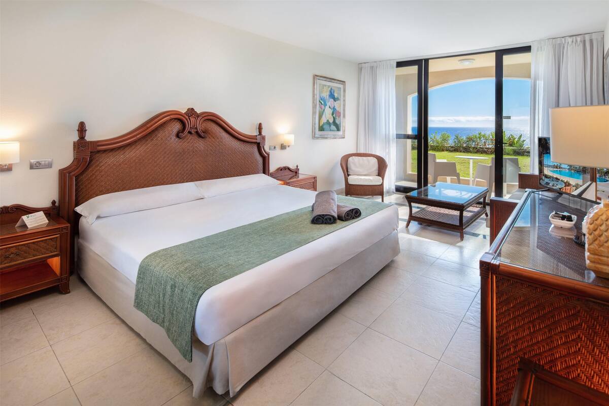 Une chambre d’hôtel avec un grand lit, un coin salon avec une table et des chaises, un fauteuil en osier et une vue sur l’océan.